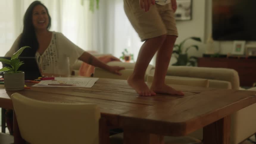 A child stomps around on a wooden table as his mother sits behind him laughing