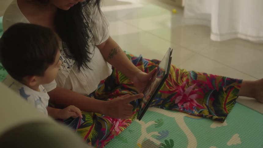 Woman with white shirt and colorful pants sits on floor with son using tablet in the living room