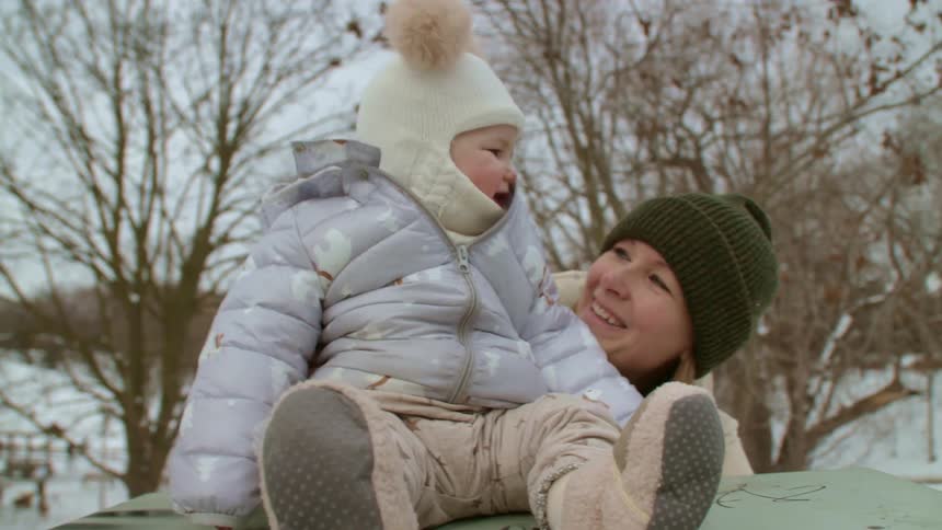 A mother laughs and plays with a sitting baby daughter outdoors in the winter