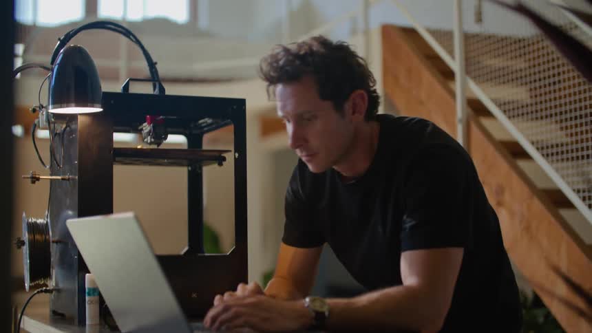 Man leans against table typing on laptop and prepping 3d printer