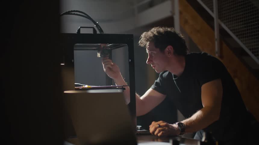 A man leaning on a desk in front of a laptop holding a piece of equipment and trying to insert it into the box next to him