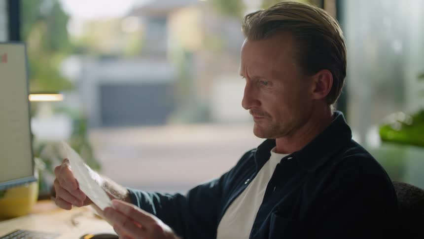A man uses his computer and reads a paper at his desk