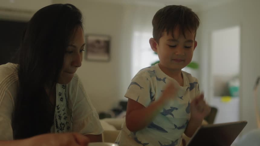 Mother and son sitting at dining table in the morning looking at computer while laughing and smiling probably home schooling