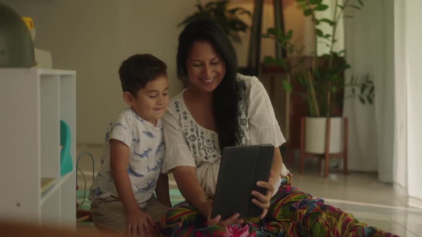 Mother with wavy hair white shirt and colorful pants shows tablet son sitting by