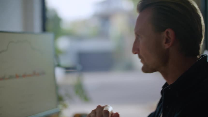 A man is sitting at a desk near a window typing on a computer keyboard as he looks at the monitor