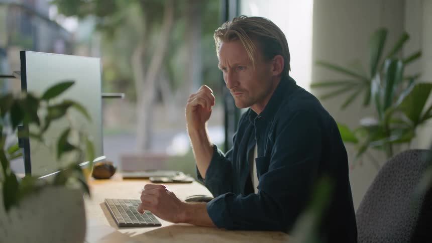 A man sitting at his desk looking at a laptop screen while typing on a computer keyboard in front of the computer screen