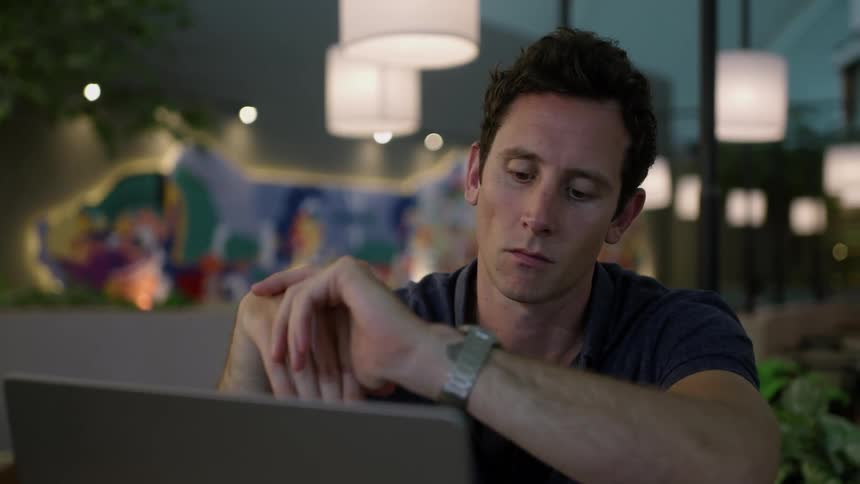 A man is sitting at a desk with a laptop computer with his hands on his face as he looks around and looks at his wristwatch