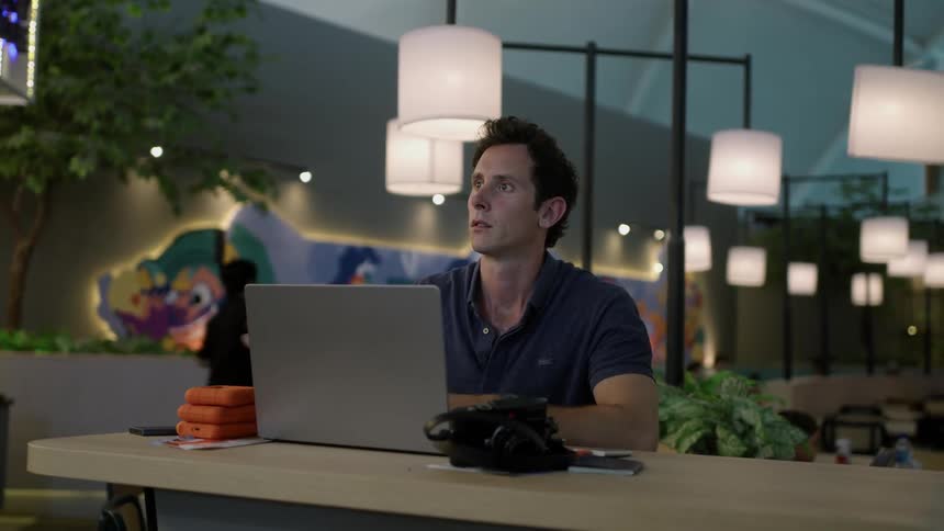 A man sitting at a table in front of his laptop looking at three orange objects and then looking at his watch