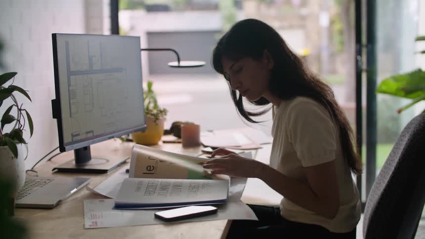 Woman sitting in office flips through book and considers it and the monitor screen contents