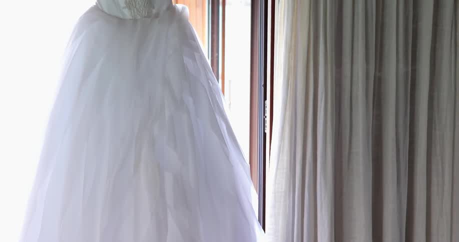 A wedding dress hangs in front of a window