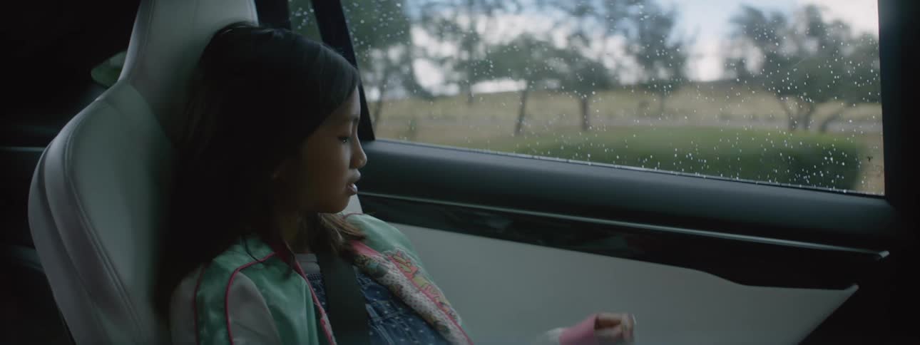 A girl rides in the backseat of a car looking out the window and smiling at someone off camera