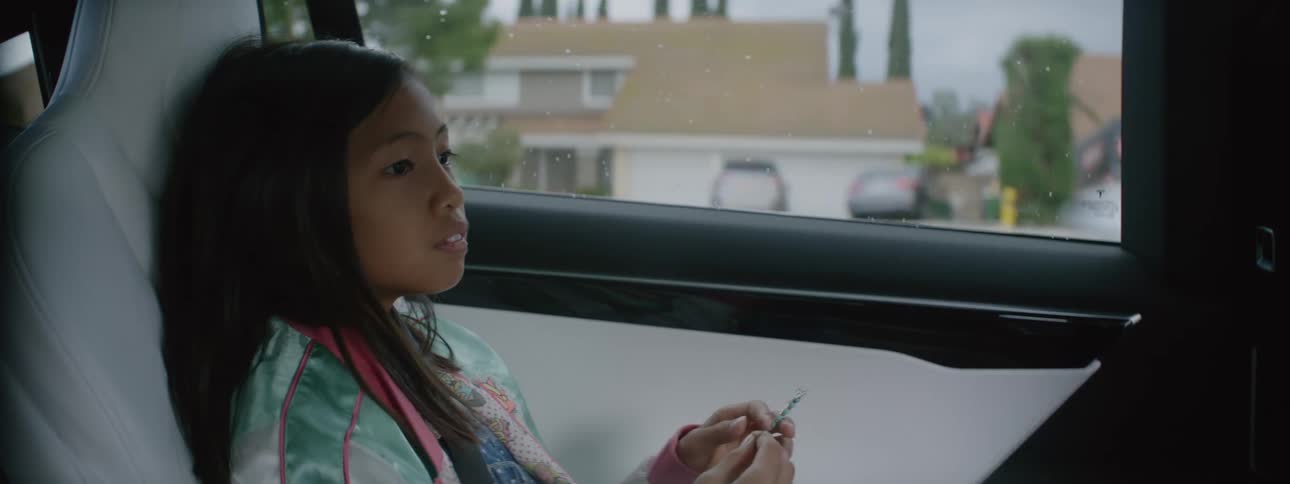 A young girl riding in the back of a car as houses and other cars go by in the window