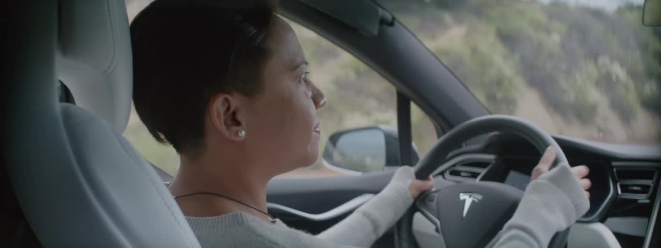 A woman is driving an automobile and she lifts her hand off the steering wheel as she talks
