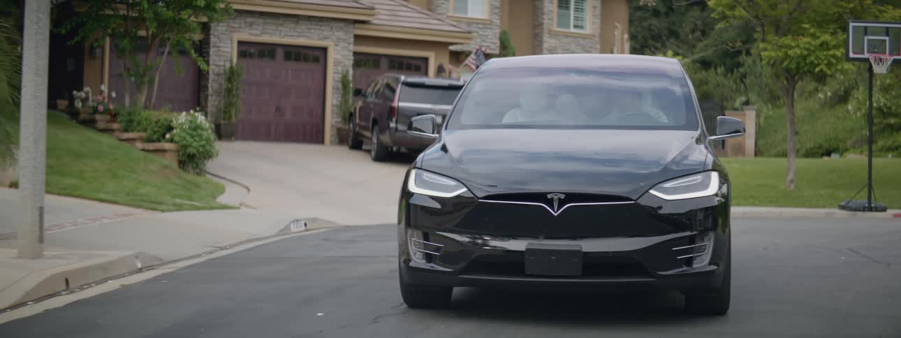 A black tesla car slowly drives through a green suburban neighborhood among homes and trees