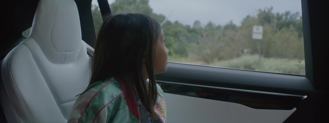 A girls is sitting in the seat of an automobile and laughing as she looks out the window at the woods