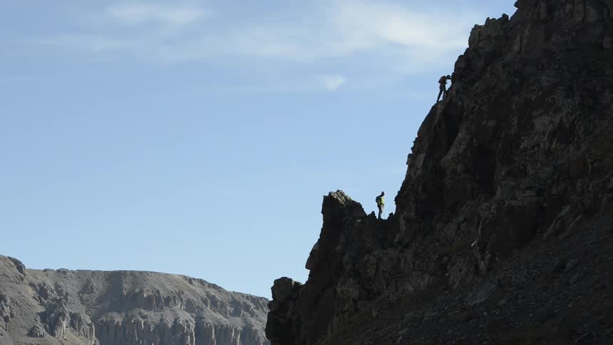 A man is standing on a mountain as he watches another man climb higher up on the ridge