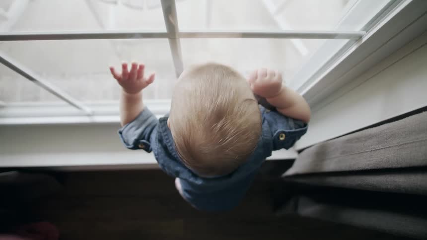 A baby looks out of a window while standing on the window sill