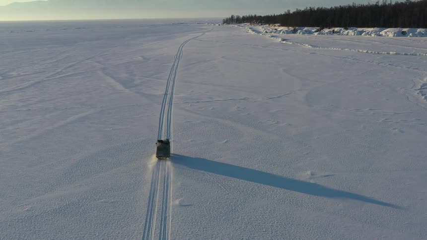 A truck drives along tire tracks in a snowy plain toward an evergreen forest at daytime