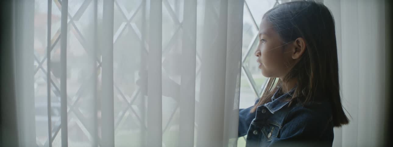 A young kid is looking outside of the window inside of there home