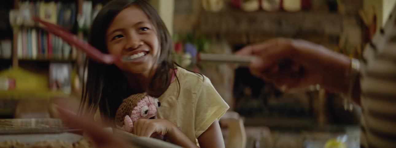 A joyful young girl holding a sock puppet in her right hand leans against the counter in an exaggerated way as she interacts with her mom who is holding a pink spatula