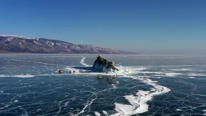 An aerial of ice covered waters with a rock formation coming out of the ice as people surround it