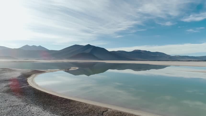 People stand on narrow strip of sand surrounded by reflective water across from mountain on cloudy day