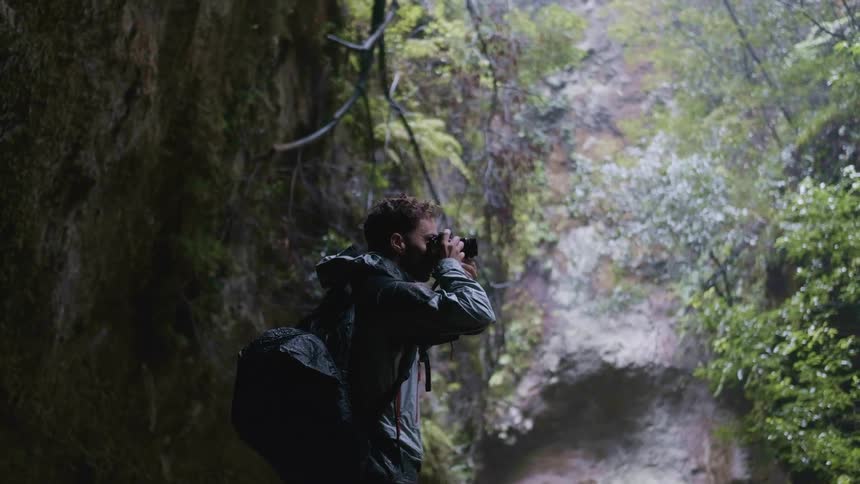 A man wearing a backpack is standing in the woods as he raises his camera to take a picture