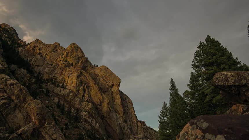A time lapse of a rocky mountain cliff and tall pine trees with clouds rolling across the sky