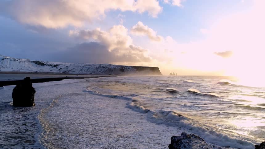 Rocks stand on the beach as waves roll in to shore and a snow covered promontory is in the distance