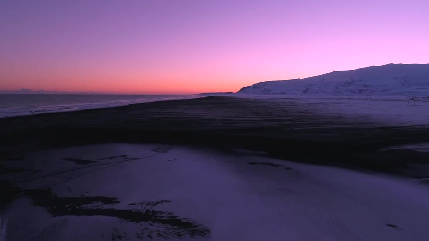 A pink and purple sunrise on the horizon with white mountain and black sand in the front