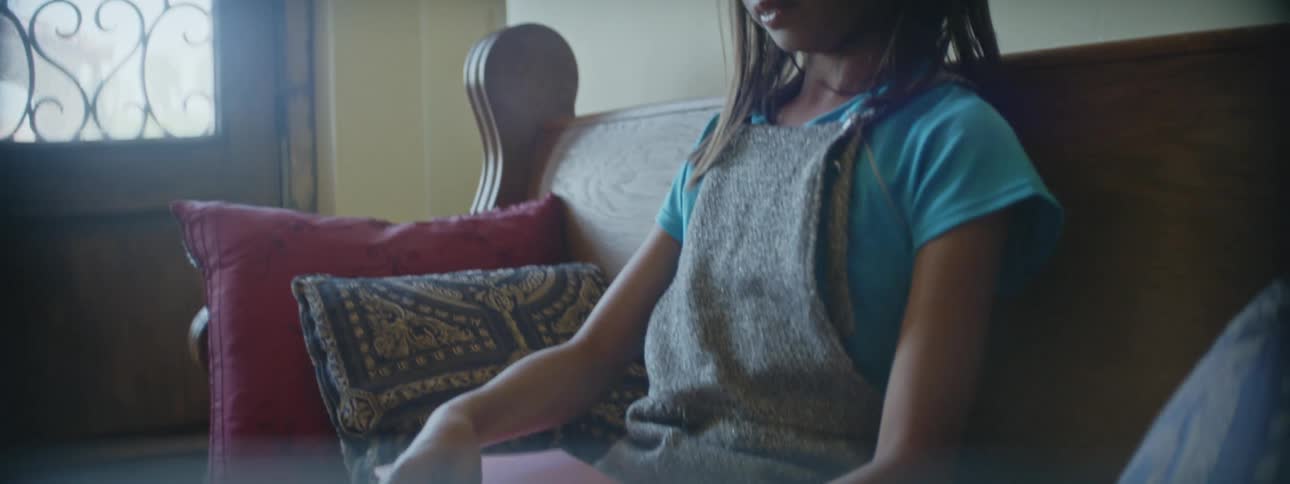 A girl uses a pen to write on a folded piece of paper while sitting on a sofa indoors