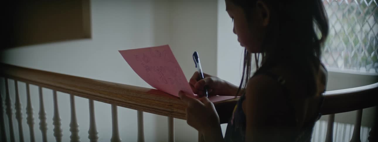 Young girl writes on a card reclined on the stairs railing at home