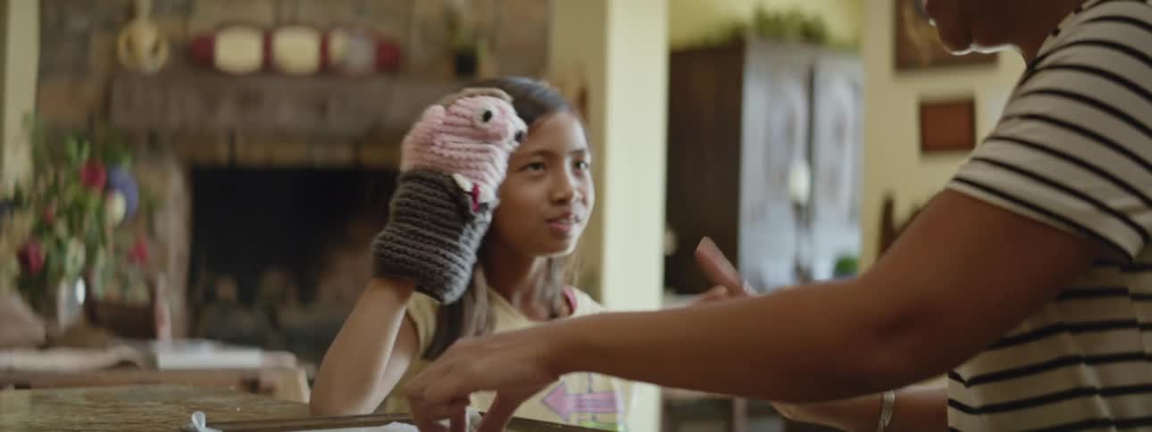 A mother and young girl standing at a kitchen counter eating cookies and laughing at the puppet on the girls hand