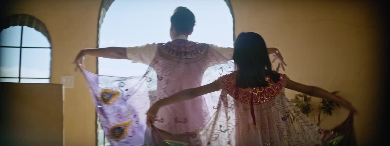 Mother and daughter spread filmy embroidered cloaks while walking toward sunny balcony