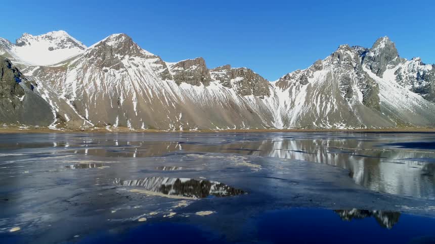 A drone view of icy waters leading up to a coastline with tall mountain ranges with snow at the peaks