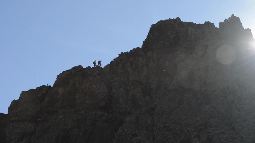Two distant hikers walk across the top of a steep rocky peak in the middle of the day