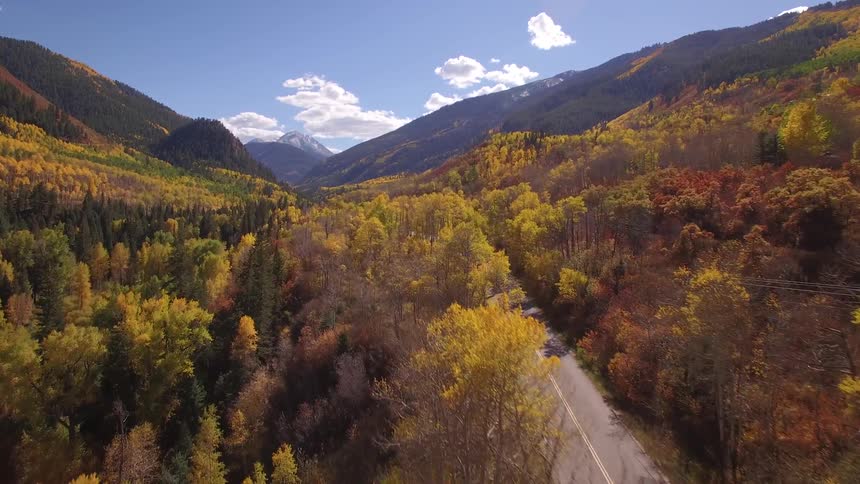 A red truck travels on a rural road surrounded by trees in beautiful autumn colors and there are mountains in the distance