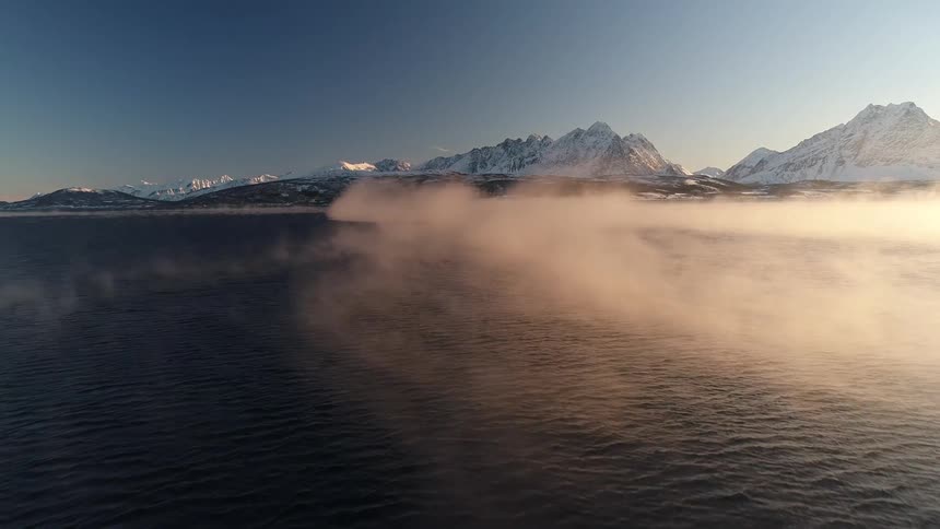 Aerial view of ocean against icy mountains or glacier ice with steam moving across the water from the sunrise