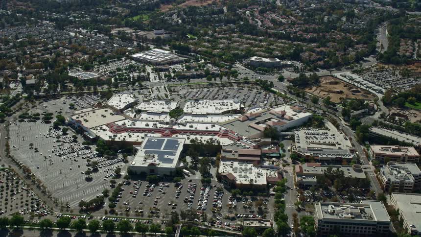 Aerial footage of Westfield Valencia Town Center a shopping mall in Santa Clarita California bustling with customers and vehicles during a partly cloudy summer day