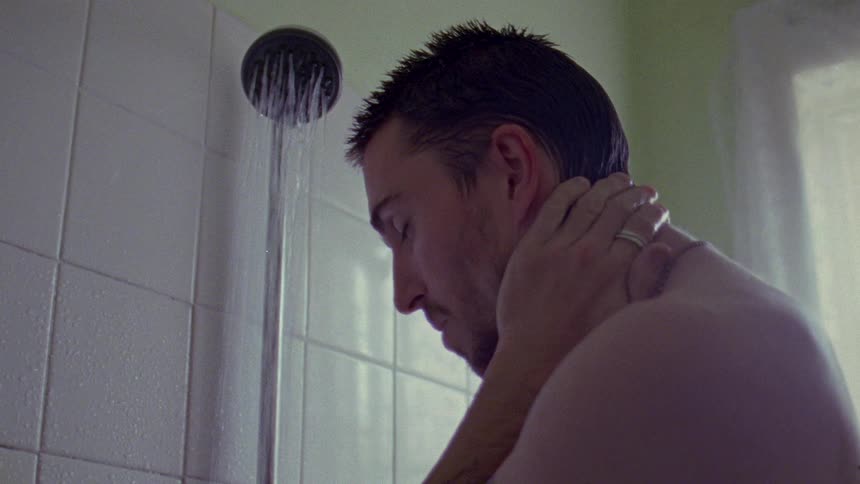 A man standing in the shower looking down as he rubs his hands across his head