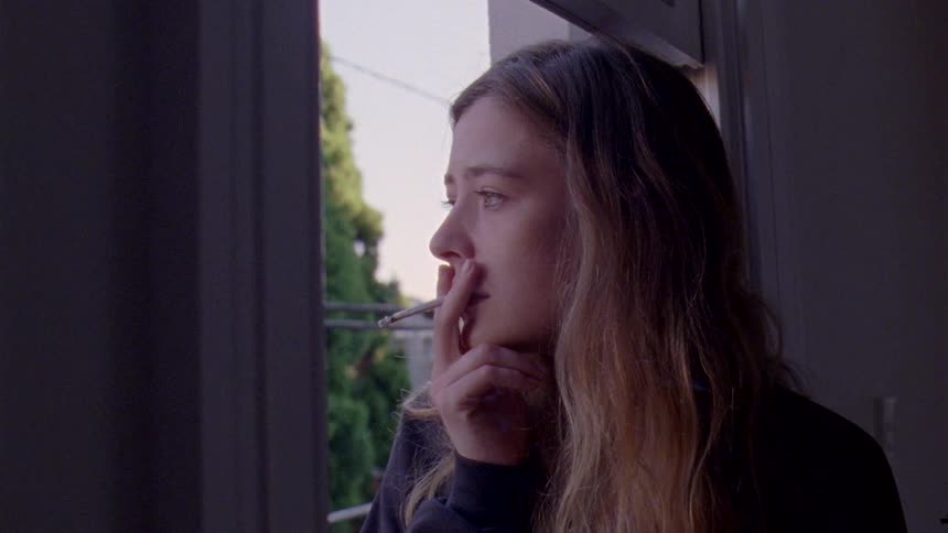 A woman standing in a window smoking a cigarette looking out the window with a sad expression