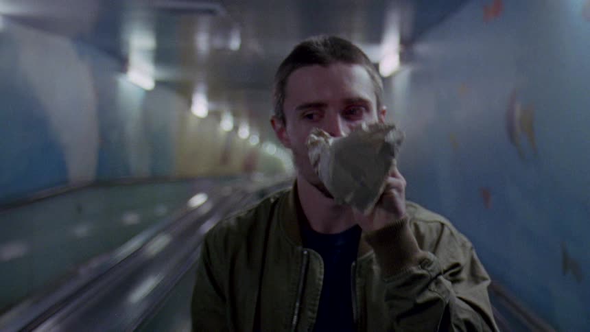 A man riding an escalator very depressed looking and drinking alcohol out of a bottle in a bag