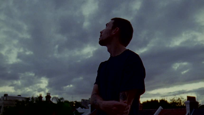 Man stands holding his arm and looking over nearby low rise building under an overcast sky