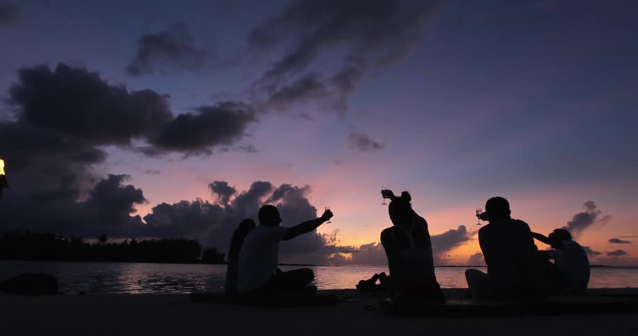 A group of people sit in a circle on the beach at sunset and raise their glasses in a toast