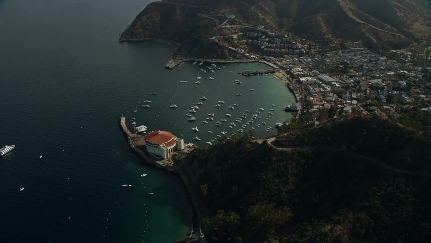 Reverse view of the harbor and coastal town of Avalon on Catalina Island California captured in stunning aerial footage during the spring or summer with partly cloudy skies and boats parked in the marina