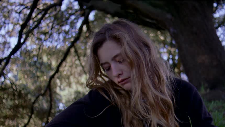A teenage girl with long blond hair is sitting outside near a tree looking sad as a man paces back and forth behind her