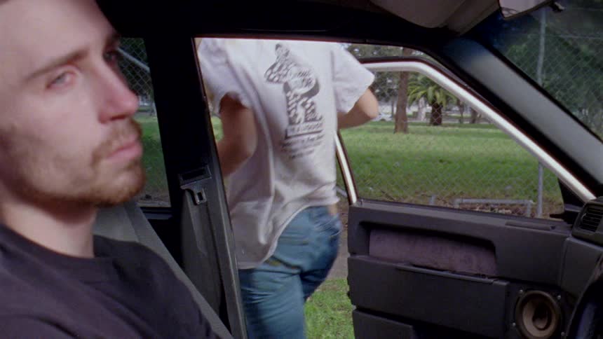 A man looking out the window of a parked car looking depressed while the woman in the driver seat gets out aggressively