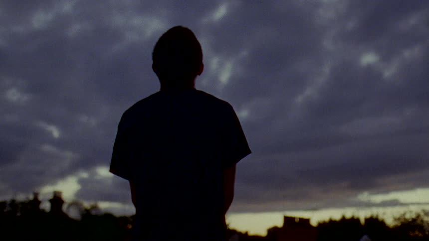 A silhouette of a man standing outside as the sky turns to dusk and clouds cover the sky