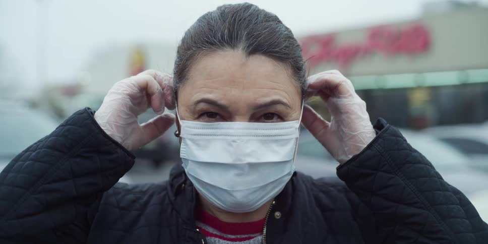 A woman already wearing gloves stands in a store parking lot and puts on a mask