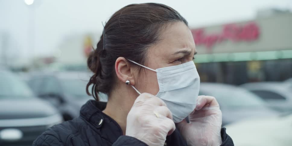 A woman wearing gloves adjusts the mask on her face before walking toward a store